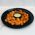 The Zeus Bowl V2 features seasoned chicken kebab pieces and roasted potato cubes arranged on a black plate around a cup of creamy dipping sauce.