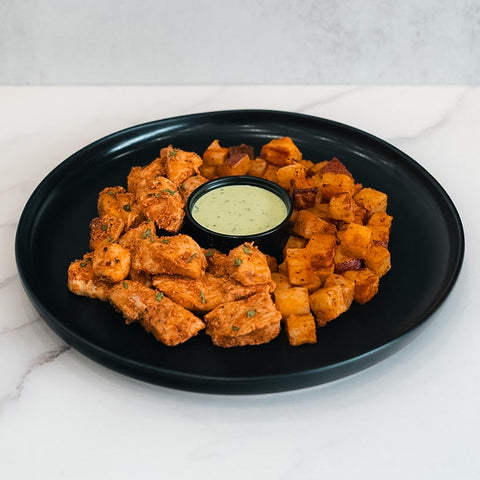 The Zeus Bowl V2 features seasoned chicken kebab pieces and roasted potato cubes arranged on a black plate around a cup of creamy dipping sauce.